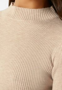 Pull en maille côtelée beige clair avec col montant, présentant de fines rayures verticales, une texture douce et une silhouette ajustée.