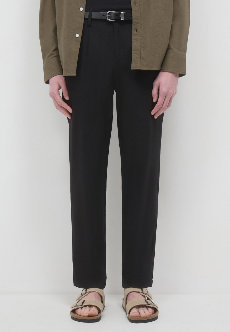 Schwarze Hosen mit maßgeschneidertem Schnitt, die eine glatte Textur haben, kombiniert mit beigen Sandalen mit verstellbaren Riemen. Olivgrüne Button-Down-Hemd.