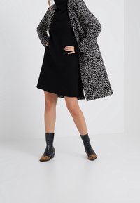Svart ribbad klänning med hög krage, lager under en grå leopardmönstrad kappa. Svarta ankbooties med ormskinnsdetaljer. Minimalistisk bakgrund.