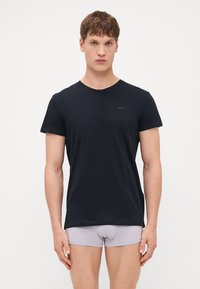 T-shirt nera a maniche corte in tessuto morbido, con scollatura rotonda e un piccolo logo del marchio sul petto. Abbinata a slip grigi chiari.