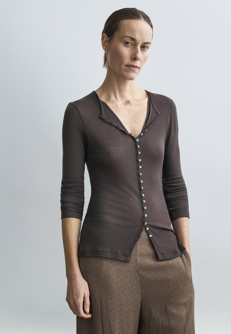 Világosbarna hajú nő, aki hátrafogott hajjal, sötétbarna, gombos, testhezálló felsőt és laza, világosbarna, texturált nadrágot visel, egyszínű háttér előtt pózol.