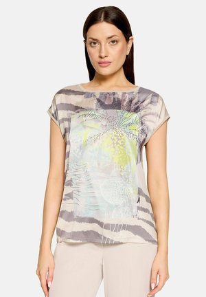 Kobieta z długimi ciemnymi włosami w krótkim rękawie, wzorzystej bluzce z abstrakcyjnymi pastelowymi i szarymi paskami oraz beżowych spodniach.