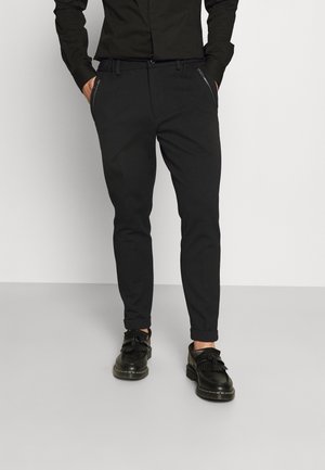 Uomo che indossa pantaloni neri slim fit con tasche con cerniera, abbinati a mocassini neri con nappine e camicia nera infilata dentro.