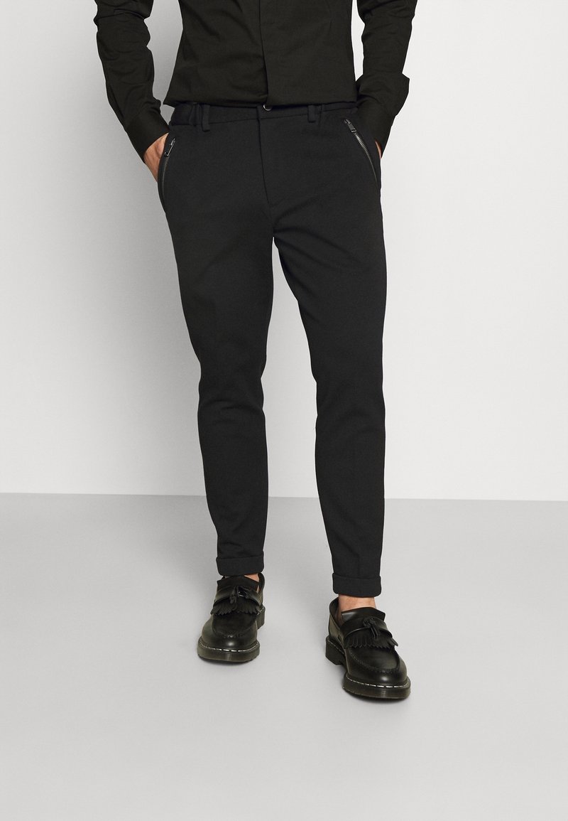 Homme portant un pantalon noir slim avec des poches zippées, associé à des mocassins noirs à pampilles et une chemise noire rentrée.