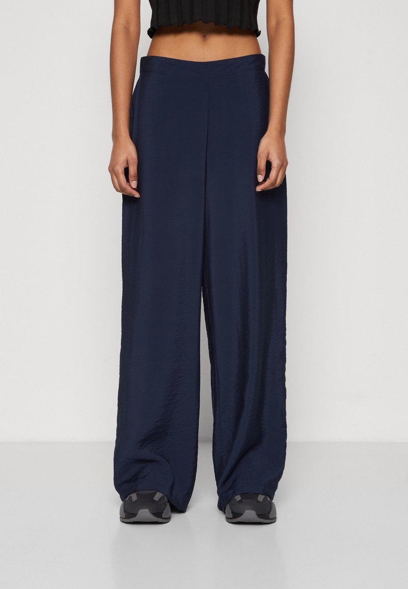 Personne portant un pantalon large bleu foncé, un haut court noir et des baskets grises et noires, debout devant un fond uni.