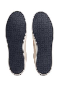 Pár světle béžových bot z pohledu podrážky, ukazující tmavé vzorované gumové podrážky s geometrickým vzorem.