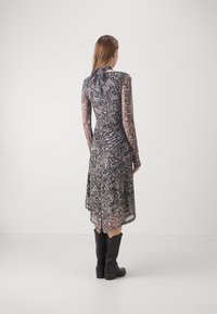 Femme portant une robe à manches longues, longueur genoux, imprimé peau de serpent, avec un dos froncé et des bottes noires mi-mollet, debout de dos sur un fond uni.