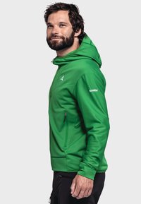 Grüne zip-up Kapuzenjacke mit texturiertem Finish, mit einem kontrastierenden Logo und seitlichem Reißverschlussfach, getragen über schwarzen Hosen.