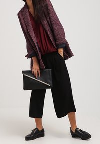 Žena v bordó blůze, texturovaném fialovém saku, černých širokých kalhotách, černých mokasínech, držící černou kabelku s ozdobným stříbrným zipem.