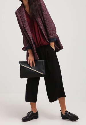 Nainen maroon-värisessä paidassa, tekstuuri-purppuravärisessä bleiserissä, mustissa leveissä housuissa, mustissa loafer-kengissä, pitäen mustaa clutch-laukkua, jossa on hopeinen vetoketju.