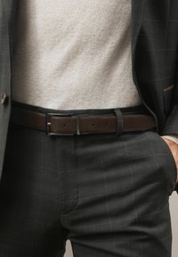 Ruskea nahkavyö, jossa on kiiltävä hope buckle, käytetty musta ruudullinen housujen päällä, yhdistetty vaaleanbeigeen tekstuuripaitaan.