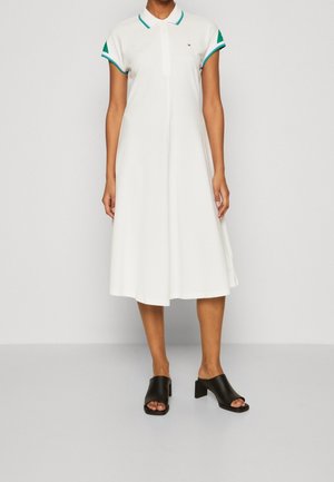 Weißes Polokleid mit kurzen Ärmeln und grünen Akzenten an Bündchen und Kragen. Ausgestelltes Design mit knielangem Saum. Schwarze offene High Heels.