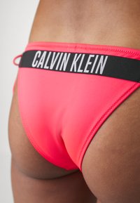 Jasne ružové bikiny s čiernym elastickým pásom s logom "CALVIN KLEIN". Hladká textúra a priliehavý strih, ktorý zdôrazňuje postavu.