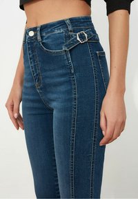 Jeans de mezclilla azul de cintura alta con un detalle de hebilla en la cinturilla, que cuentan con una textura suave y sutiles acentos de costura.