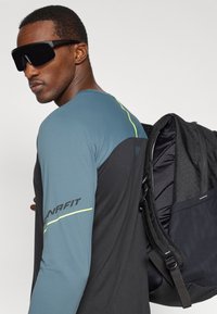 Maglietta nera a maniche lunghe con pannello turchese, accenti gialli e logo; indossa occhiali da sole neri di grandi dimensioni; porta uno zaino nero con superficie testurizzata.