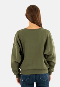 Sudadera de gran tamaño en verde oliva con hombros caídos, puños y dobladillo de canalé, fabricada en una tela suave. Llevada con jeans de mezclilla azules.