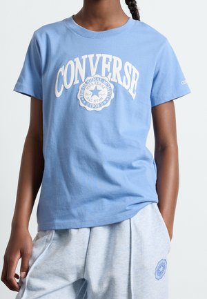 Enfant portant un t-shirt Converse bleu clair avec logo et un pantalon gris clair, debout avec une main dans la poche.