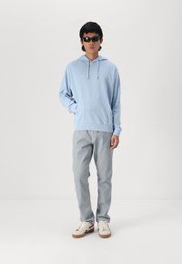 Felpa azzurra con cappuccio e tasca frontale, abbinata a pantaloni grigi chiari. Il modello indossa occhiali da sole neri e sneakers bianche.