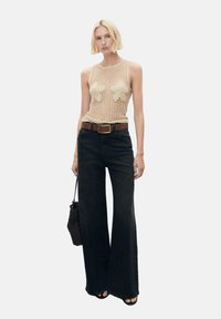 Beige knitted tank top with floral accents, paired with high-waisted black flared jeans and a brown belt; model holding a black bag.