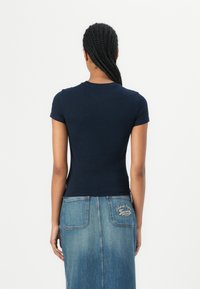 T-shirt in cotone blu navy, aderente, con maniche corte e scollo rotondo. Il retro presenta un piccolo logo e dettagli di cucitura. Indossato con una gonna di jeans.