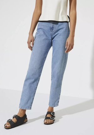Mujer que lleva pantalones vaqueros azul claro de talle alto, camiseta sin mangas blanca con ribete negro y sandalias negras con doble hebilla, de pie sobre suelo blanco.