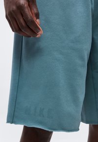 Σκούρο χέρι που ακουμπά κοντά στο γόνατο, φοράει πετρόλ σορτς με ανεπαίσθητη εκτύπωση "DUKE" κοντά στο τελείωμα, σε απλό φόντο.