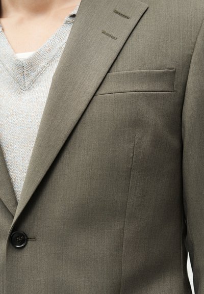 Κοντινό πλάνο ενός ατόμου που φοράει σακάκι ταμπά πάνω από μπεζ πλεκτό πουλόβερ, με λευκό εσώρουχο να φαίνεται στο λαιμό.