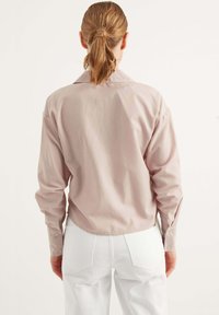 Camicia rosa chiaro a maniche lunghe con yoke posteriore, colletto classico, polsini con bottone e orlo curvo, abbinata a jeans bianchi a vita alta.