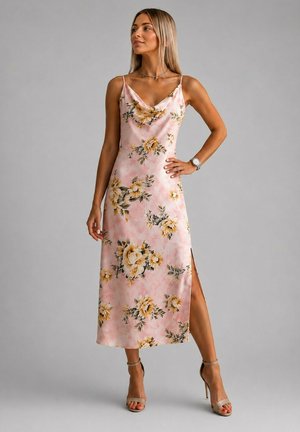 Femme portant une robe midi sans manches à fleurs roses avec une fente latérale et des sandales à talons hauts beige sur un fond gris uni.