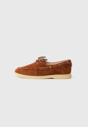 Brown suede lace-up boat shoes with a rounded toe, tan rubber sole, metal eyelets, and decorative stitching along the upper.