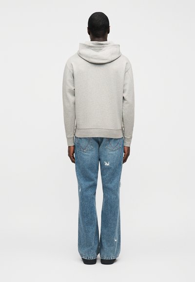 Šviesiai pilkas džemperis su gaubtu ir nusidėvėjusios mėlynos džinsinės kelnės; laisvo silueto su gofruotu apačios kraštu ir kišenėmis, matomi siūlės ir trupinimų detalės.