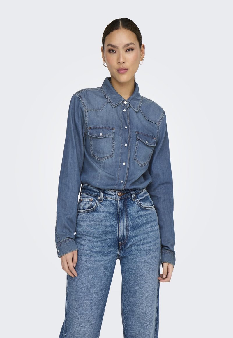 Camisa de mezclilla en azul medio, fabricada en tejido de peso medio. Cuenta con un collar, dos bolsillos en el pecho y cierre de botones, combinada con jeans de tiro alto en azul.