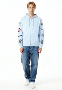 Felpa con cappuccio azzurro chiaro con cerniera, con testo grafico e immagini sulle maniche, abbinata a jeans blu larghi e scarpe da ginnastica bianche.