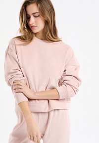Suéter rosa claro acanalado con un corte holgado, que presenta un escote redondo y mangas largas, mostrado con los brazos cruzados.