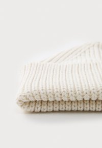 Caciula tricotată crem cu margine pliată, având o textură ribbită. Materialul pare moale și pliabil, potrivit pentru căldură și confort.