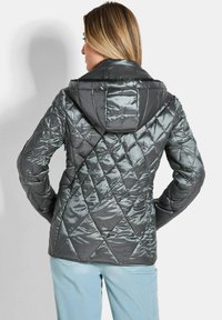 Gequilted pufferjack in donkergrijs met een glanzende afwerking, voorzien van een capuchon en diamantvormige stiksels, gecombineerd met lichtblauwe broek.
