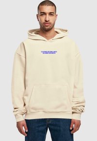 Felpa beige con tasca davanti, con testo blu: "SUPERANDO I LIMITI DEFINENDO IL CORAGGIO." Realizzata in materiale morbido con vestibilità rilassata.