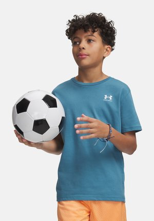 Zēns ar cirtainiem matiem, kas valkā zilu kreklu un oranžas šortus, tur melnbaltu futbolbumbu, skatoties nedaudz uz augšu un pa labi.