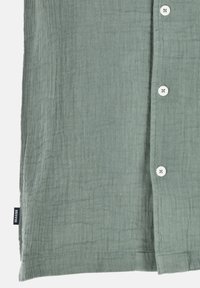 Chemise boutonnée verte à texture avec six boutons blancs et une étiquette portant la mention "SEASIDE" sur l'ourlet. Tissu léger avec une finition froissée.