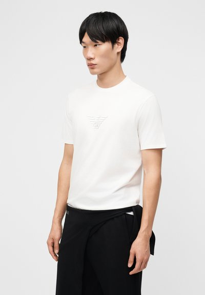 Νεαρός άνδρας με μαύρα μαλλιά που φοράει λευκό T-shirt με κοντά μανίκια και ανάγλυφο λογότυπο, καθώς και μαύρο παντελόνι με δέσμες σακάκι γύρω από τη μέση.