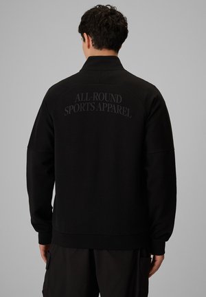 Mężczyzna w czarnej kurtce z długim rękawem z nadrukiem "ALL-ROUND SPORTS APPAREL" na górnej części pleców, odwrócony tyłem do kamery.