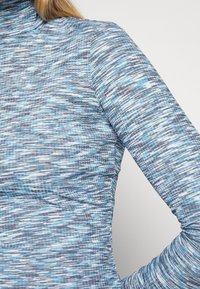 Maglia a maniche lunghe realizzata in morbido tessuto blu testurizzato con un motivo a onde in diverse sfumature di blu e grigio. Presenta un colletto alto.