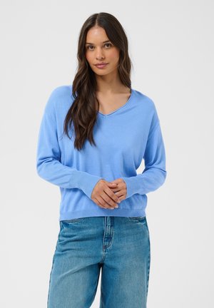 Femme aux longs cheveux foncés portant un pull bleu clair à col en V et un jean bleu, debout, les mains doucement jointes devant un fond gris uni.