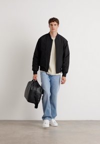 Black bomber jacket, cream sweater, light blue jeans, and white sneakers. Carrying a black bag. Neutral background, relaxed fit.