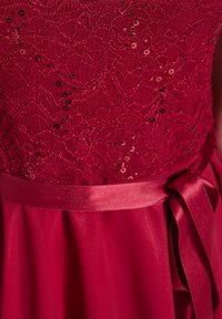 Robe rouge avec un corsage en dentelle orné de sequins. Présente un ruban en satin à la taille et une jupe fluide en tissu doux.