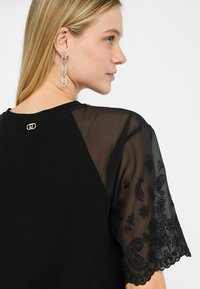 Top nero con maniche trasparenti ricamate, caratterizzato da motivi paisley e floreali. Dettaglio in metallo dorato sul retro del collo.