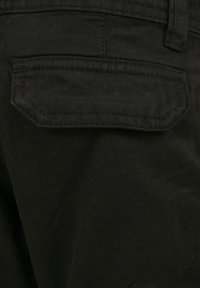 Nära bild av svart byxfabrics som visar en bakficka med lock och synlig sömnadsdetalj.