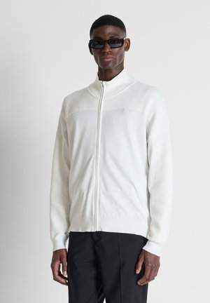 Uomo con occhiali da sole neri, maglione bianco con zip e pantaloni neri, in piedi davanti a uno sfondo chiaro e uniforme.