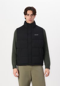 Black puffer vest with high collar, two front pockets, and subtle logo detailing. Worn over a green sweater, with a smooth texture.