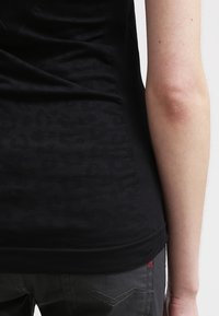 Personne portant une chemise noire ajustée et un jean gris foncé, vue de dos, mettant l'accent sur le bras droit et le côté du torse.